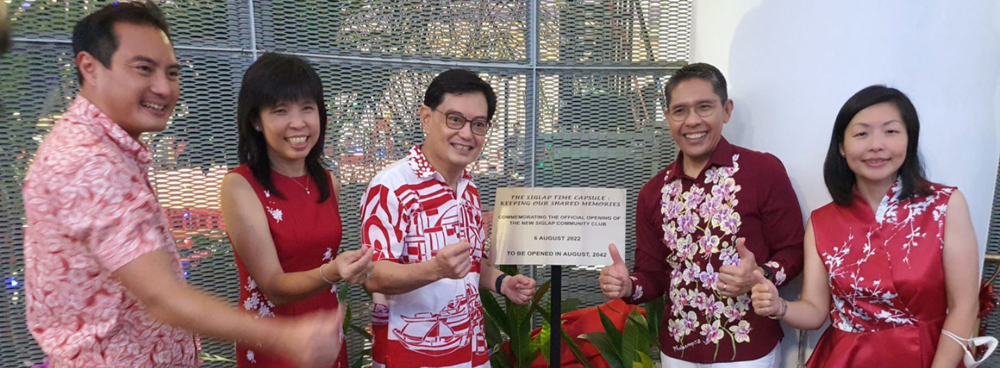 Five people wearing red and white patterned shirts and dresses pose by a plaque for the "Siglap Time Capsule".
